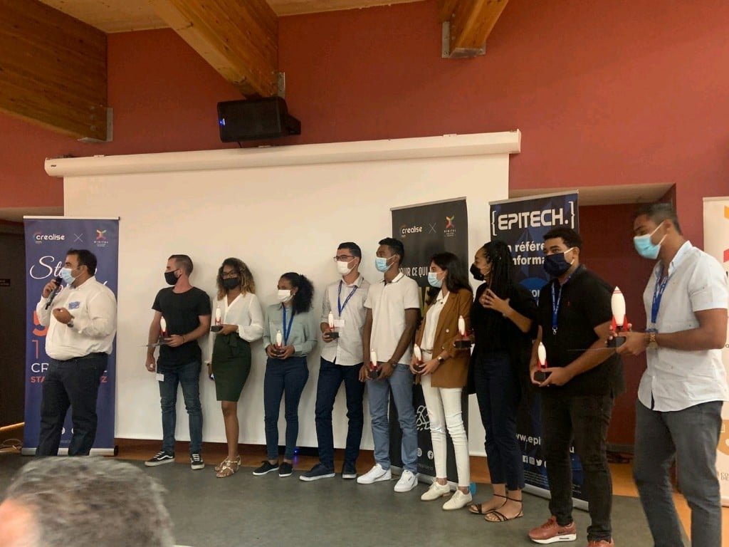 Groupe de personnes masquées tenant des trophées, debout devant des bannières d'événement.