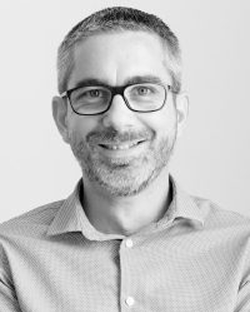 Homme souriant avec lunettes et barbe, chemise &agrave; carreaux, en noir et blanc.