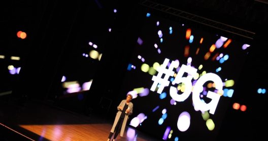 Une personne présente sur scène devant un écran affichant "#5G".